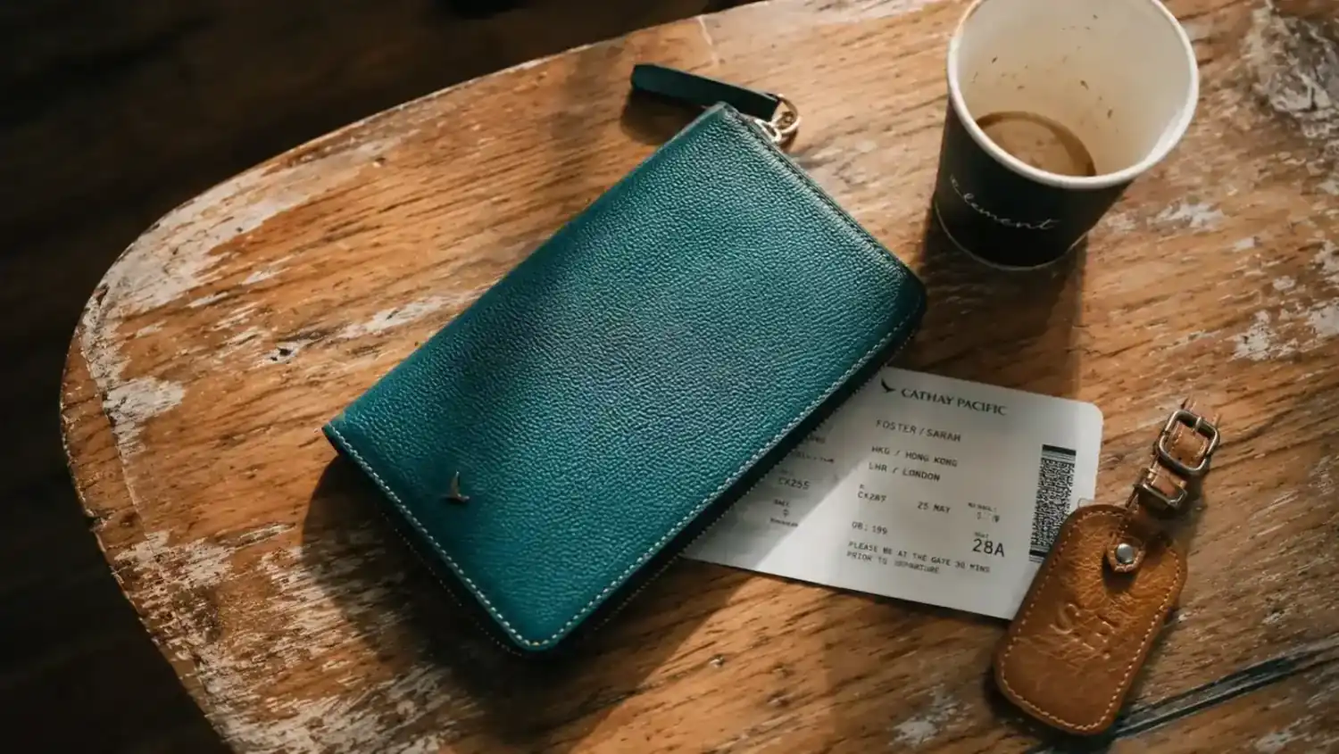 A teal RFID passport holder resting on a wooden table next to a boarding pass and a cup of coffee before a morning flight