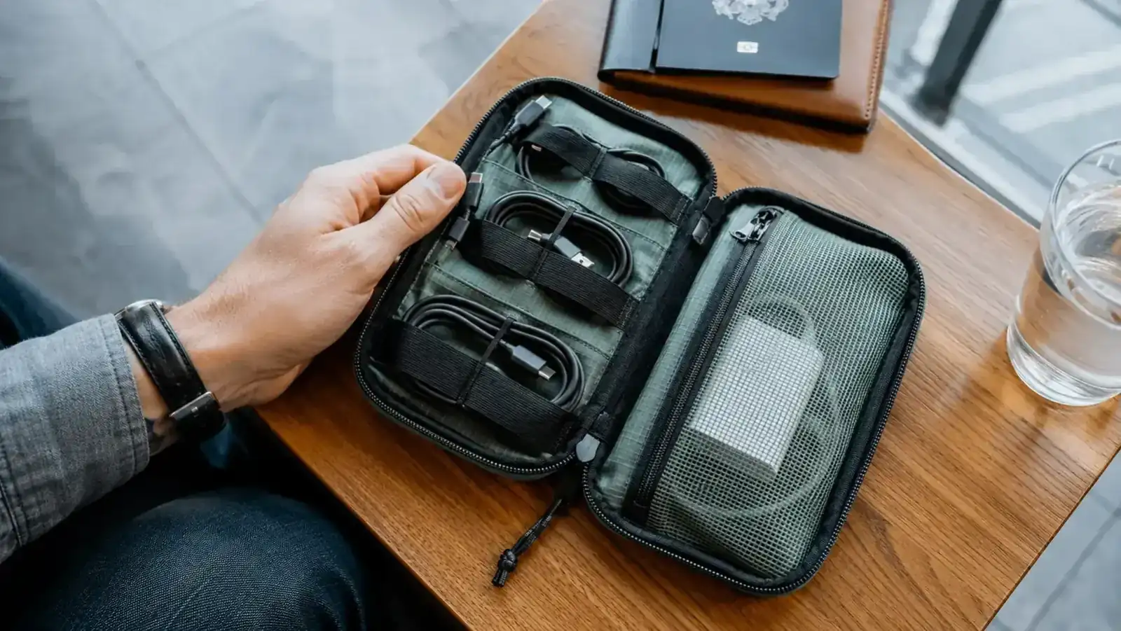 Open travel cable organizer pouch laid flat on a wooden airport lounge table, showing neatly arranged cables, USB chargers, and earbuds in elastic loops