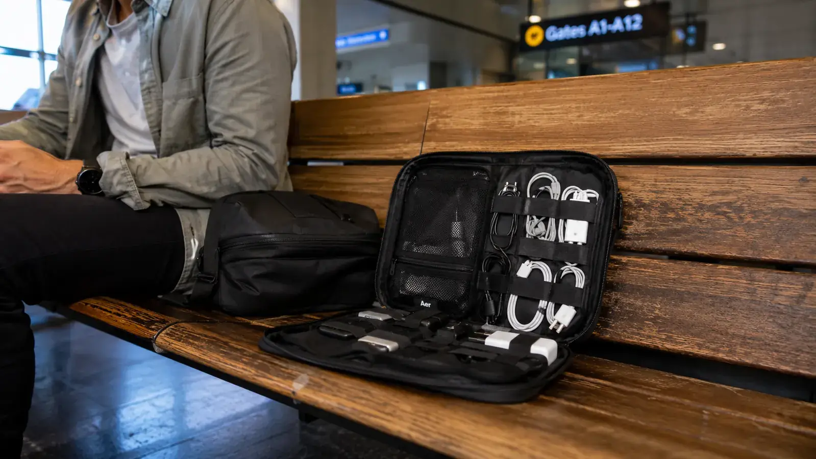 FYY electronic cable organizer bag open on a wooden airport bench showing organized cables, chargers, and earbuds