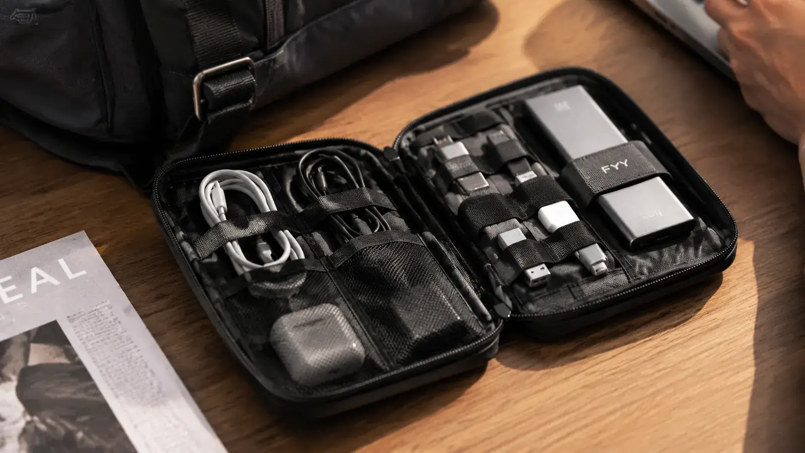 Open FYY cable organizer pouch showing neatly arranged charging cables, USB adapters, and earbuds on a wooden desk next to a laptop
