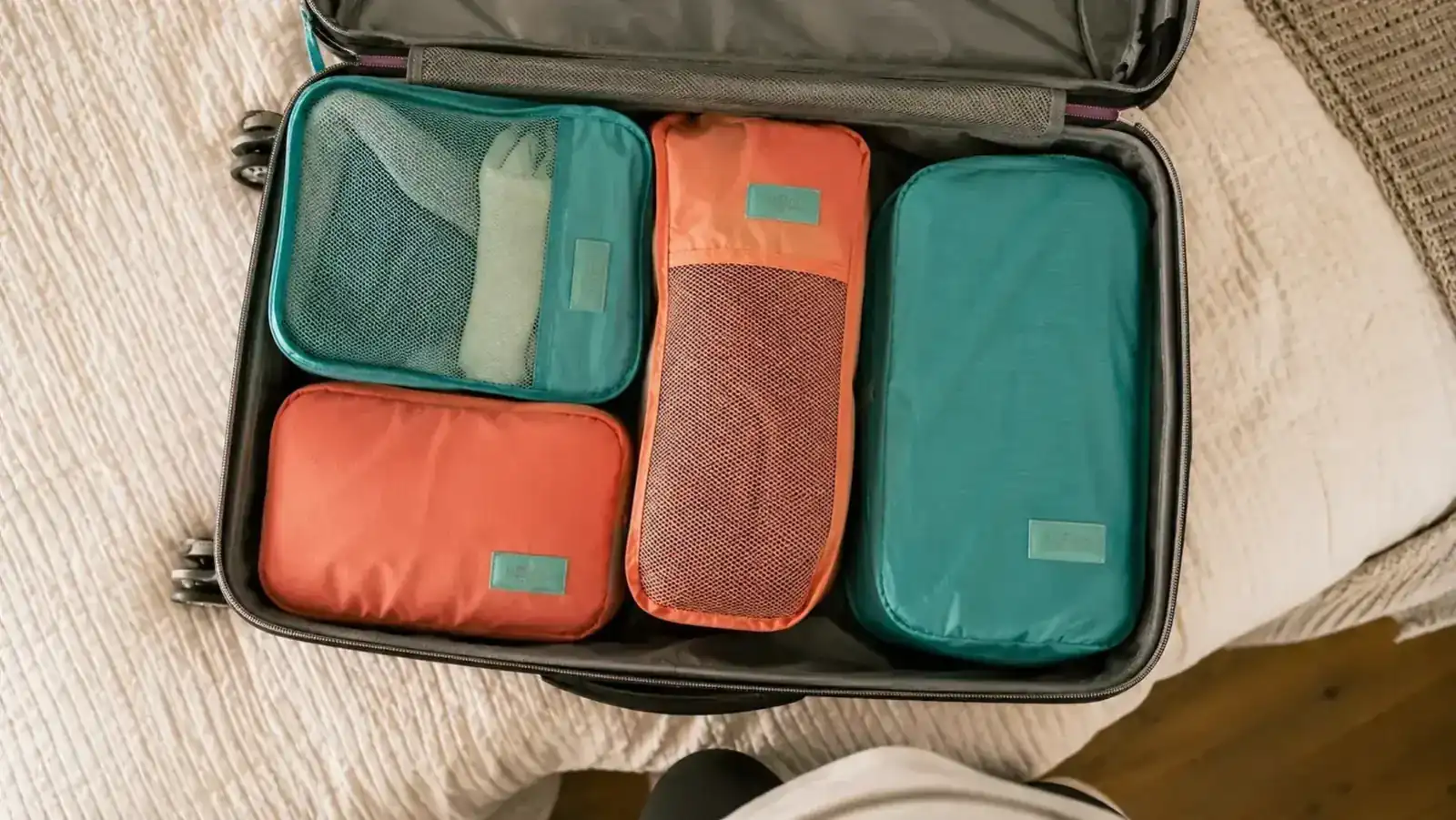 Open carry-on suitcase neatly packed with color-coded compression cubes laid out flat, bed visible in background
