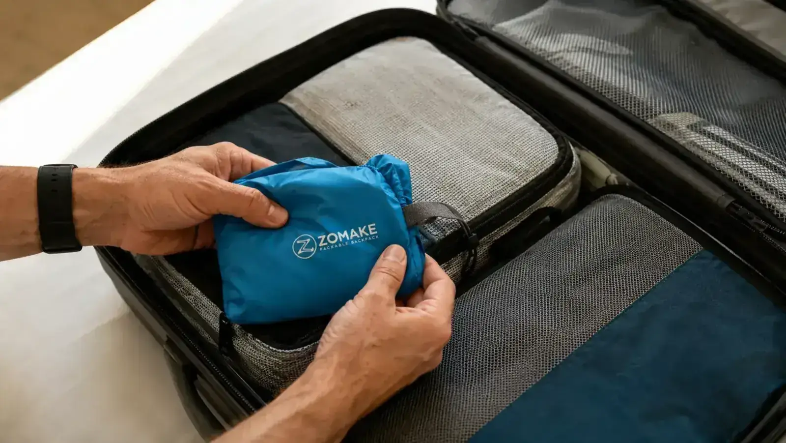 ZOMAKE packable daypack being stuffed into a small pouch the size of a fist, sitting on top of a folded carry-on suitcase
