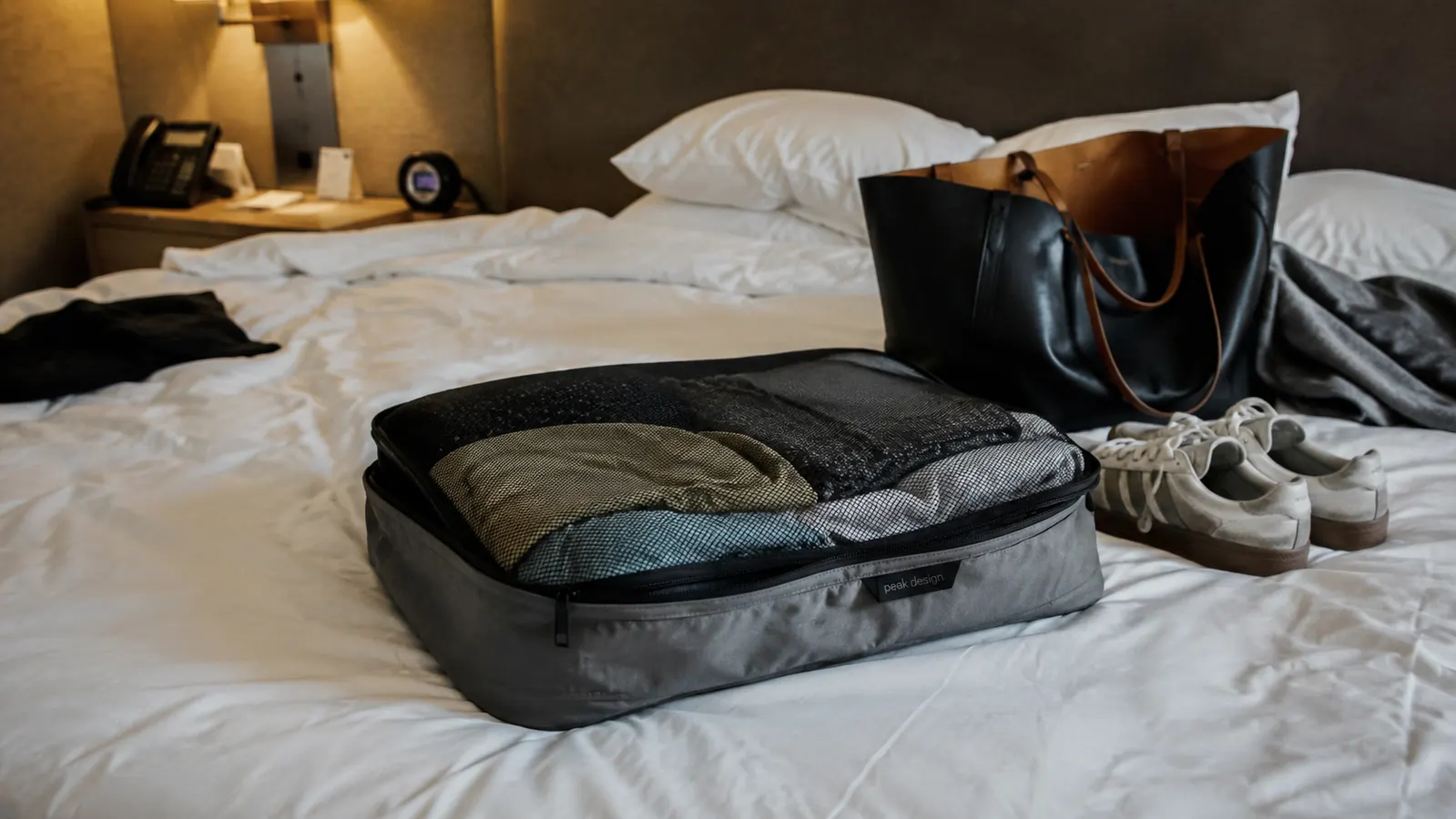 Compression packing cube sitting half-open on a hotel bed showing clothes visible through mesh top, with a tote bag and shoes nearby on a rumpled white duvet
