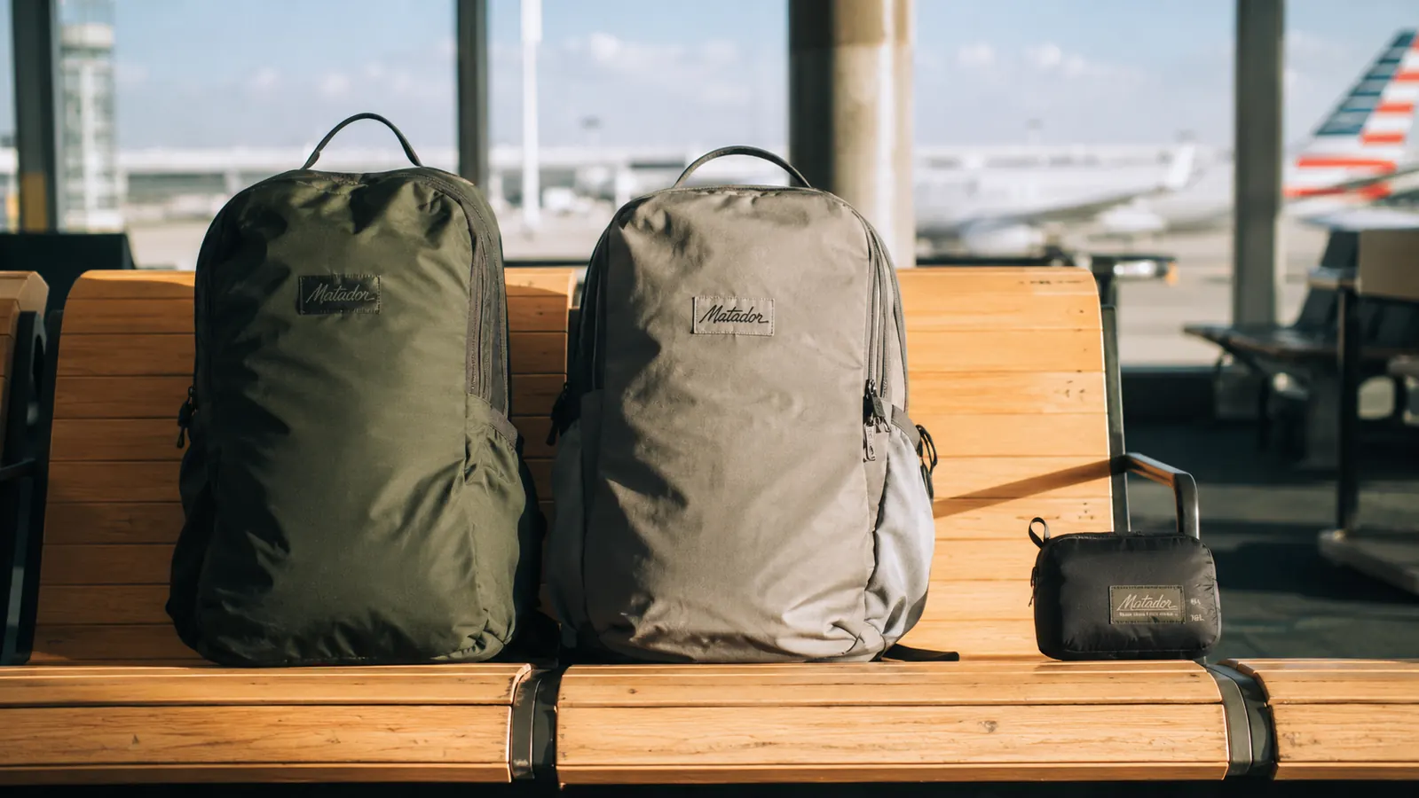 Two packable daypacks laid side by side on a wooden airport bench, one collapsed into a pouch, the other open