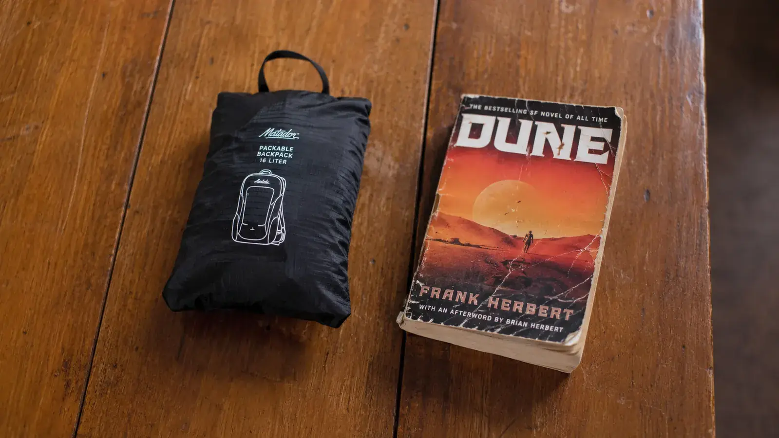 A packable daypack folded flat into its own pocket, shown next to a standard paperback book for scale