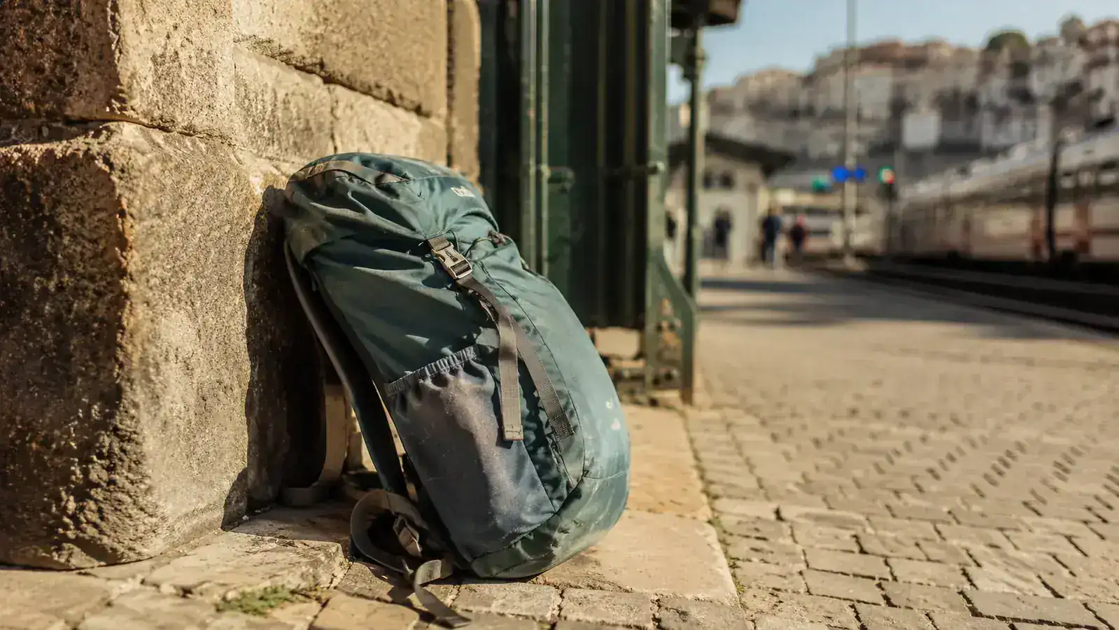 A well-worn packable daypack resting against a stone wall at a European train station, golden afternoon light